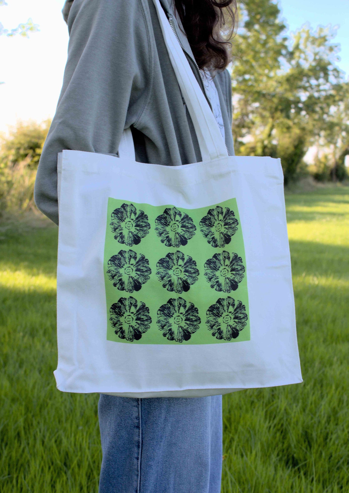 A person wearing a grey hoodie and blue jeans, carrying a white canvas tote bag with a bright green square print containing nine repeating black smiling flowers design. The person is standing in a grassy outdoor setting with trees in the background