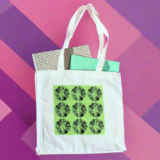 A white canvas tote bag with white handles, featuring a bright green square print with nine repeating black smiling flower designs. The bag holds a patterned box and a light blue box, set against a background of diagonal stripes in shades of purple and pink