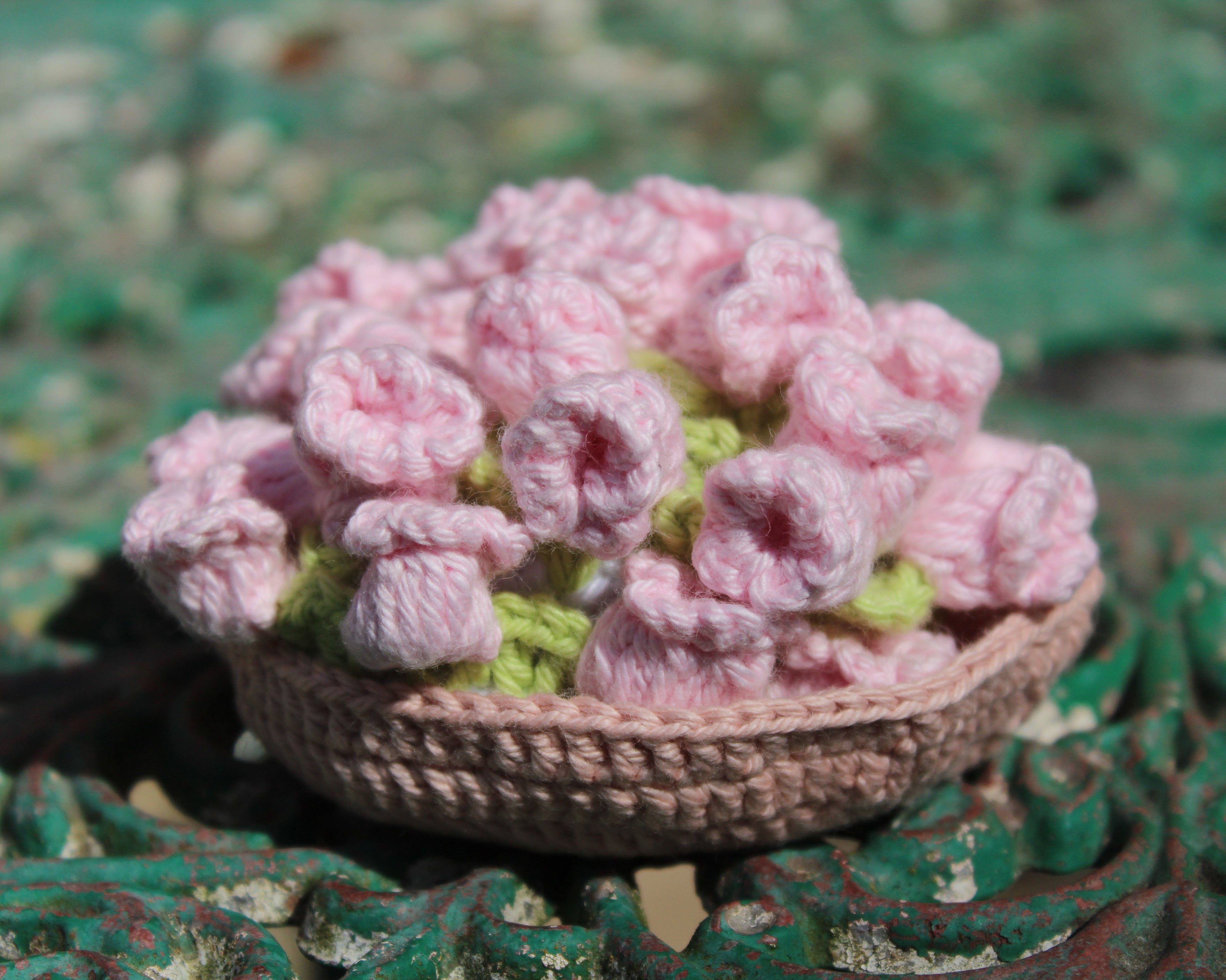 A handmade crocheted basket filled with pink yarn flowers and green yarn leaves, resting on a distressed green metal surface.