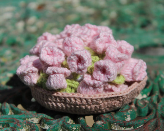 A handmade crocheted basket filled with pink yarn flowers and green yarn leaves, resting on a distressed green metal surface.