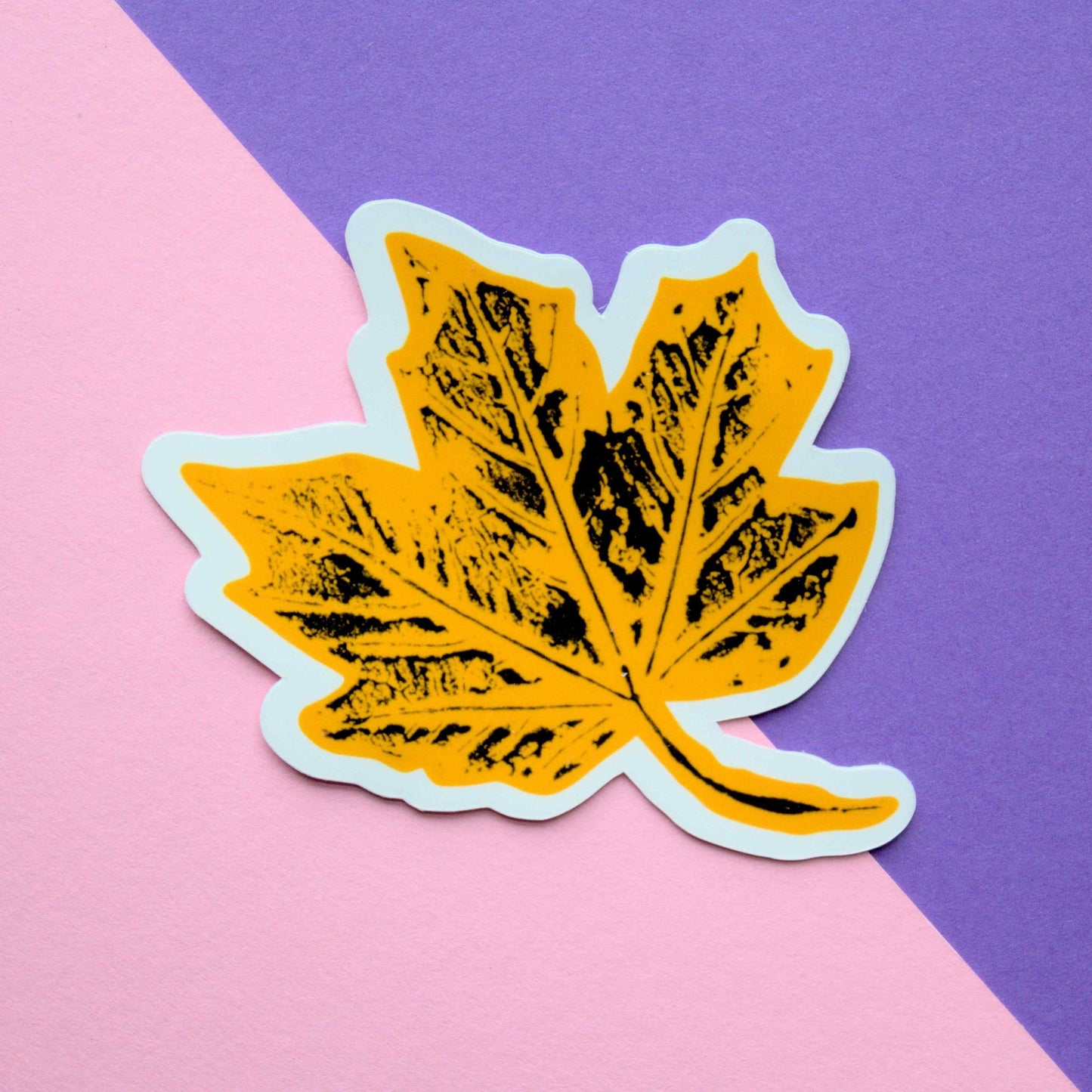 A stylized yellow maple leaf sticker with a white outline and black ink texture, placed on a background diagonally split between light pink and purple