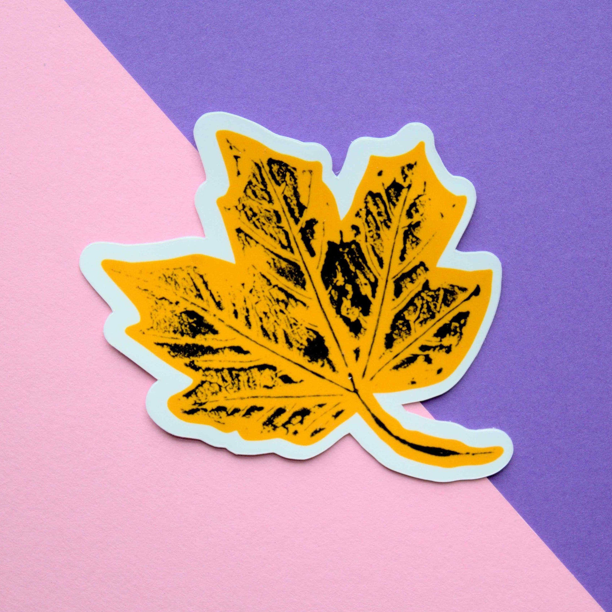 A stylized yellow maple leaf sticker with a white outline and black ink texture, placed on a background diagonally split between light pink and purple