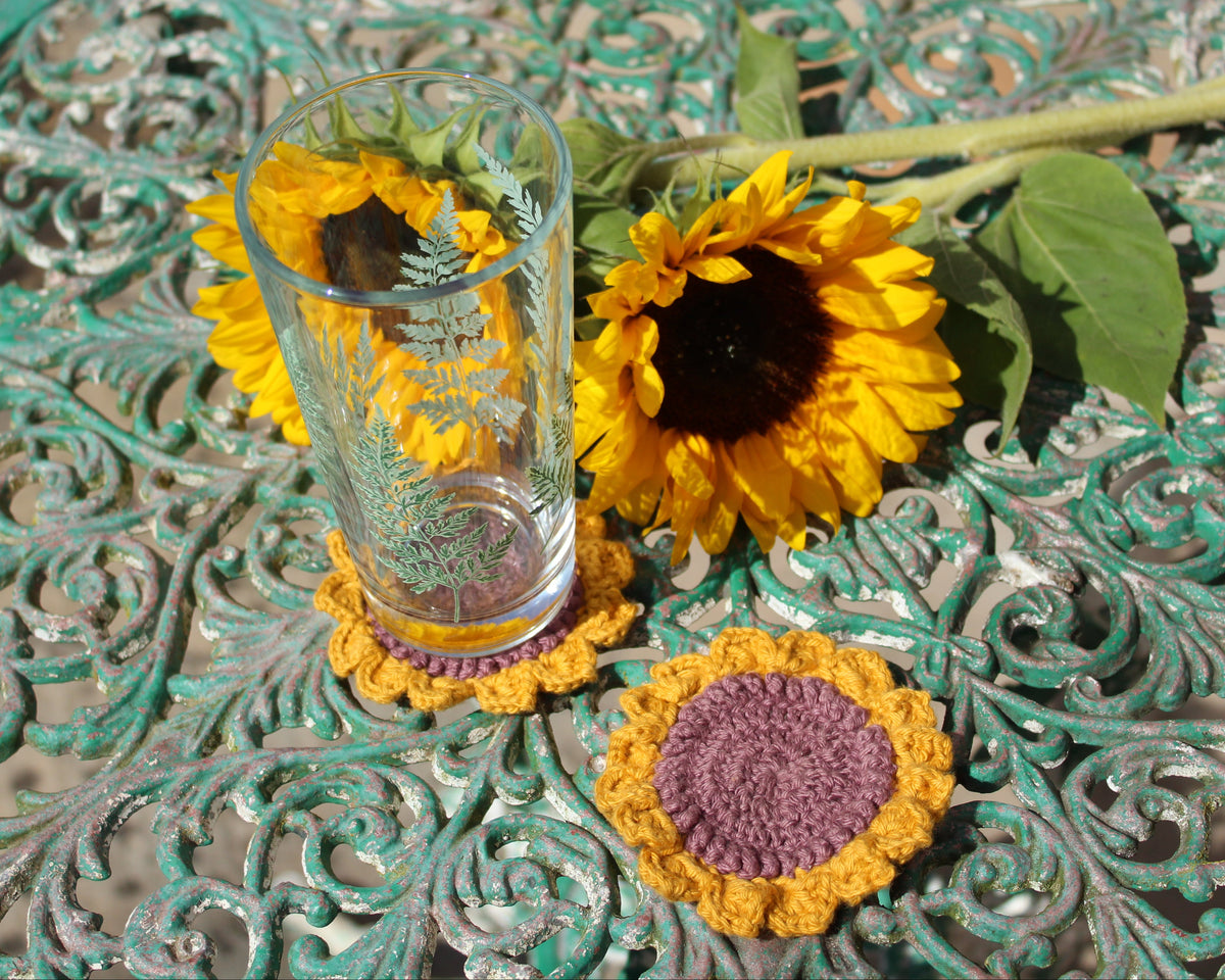 A clear glass tumbler with a fern pattern sits on a yellow and purple crocheted coaster, styled on a distressed teal metal table with sunflowers.
