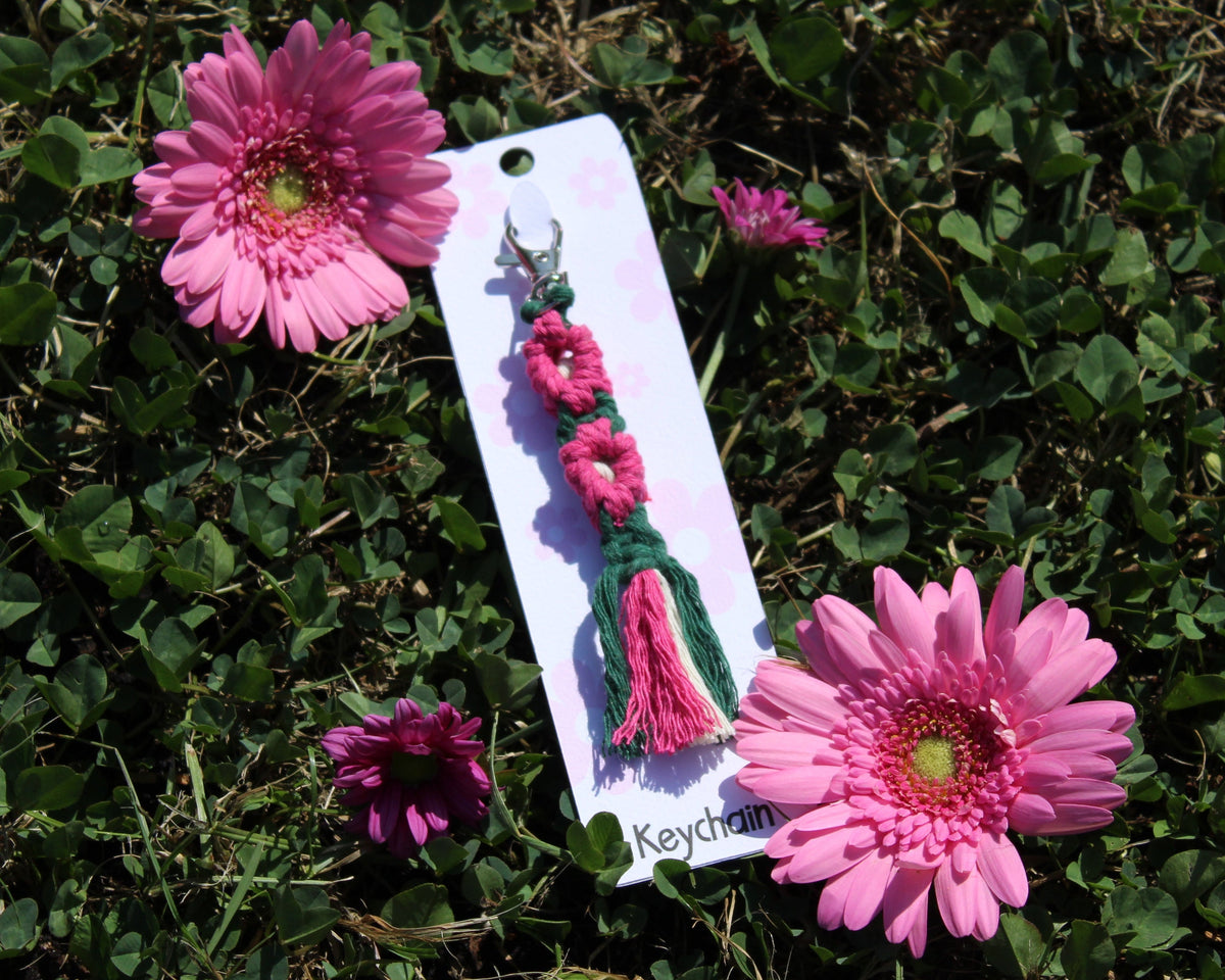 A handmade pink and green woven keychain with a tassel, attached to a white display card, placed on green grass between two pink daisy flowers.