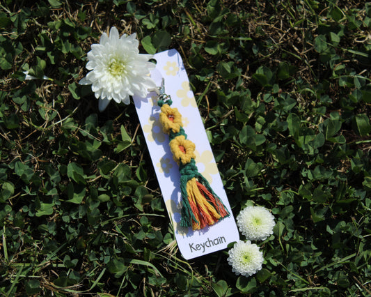 A handmade multi colour keychain with yellow flower charms and green, yellow, and orange tassels, attached to a white card labeled 'Keychain' and placed on green grass with white flowers.