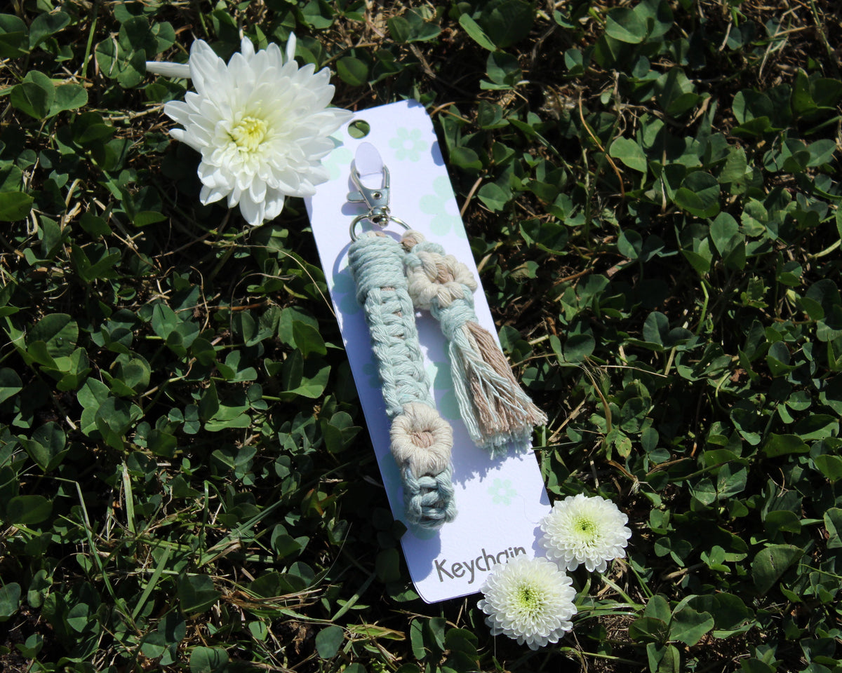 A handmade macrame keychain with a silver clasp, featuring a mint green braided cord, a beige tassel, and a small cream flower charm, displayed on its white backing card labeled 'Keychrom' and set against a background of green clover and white flowers.
