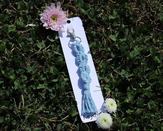 A light blue knotted tassel keychain with a silver metal clasp, displayed on a white card over a green clover background, with a pink flower and two small white flowers placed around it.