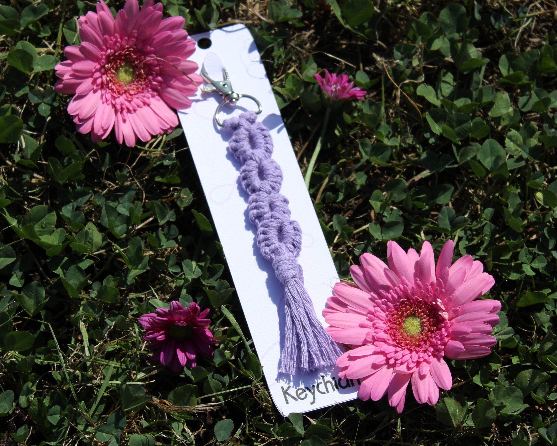 A lavender purple macrame keychain with a tassel and metal keyring, attached to a white display card, placed on green grass between two pink gerbera daisies.