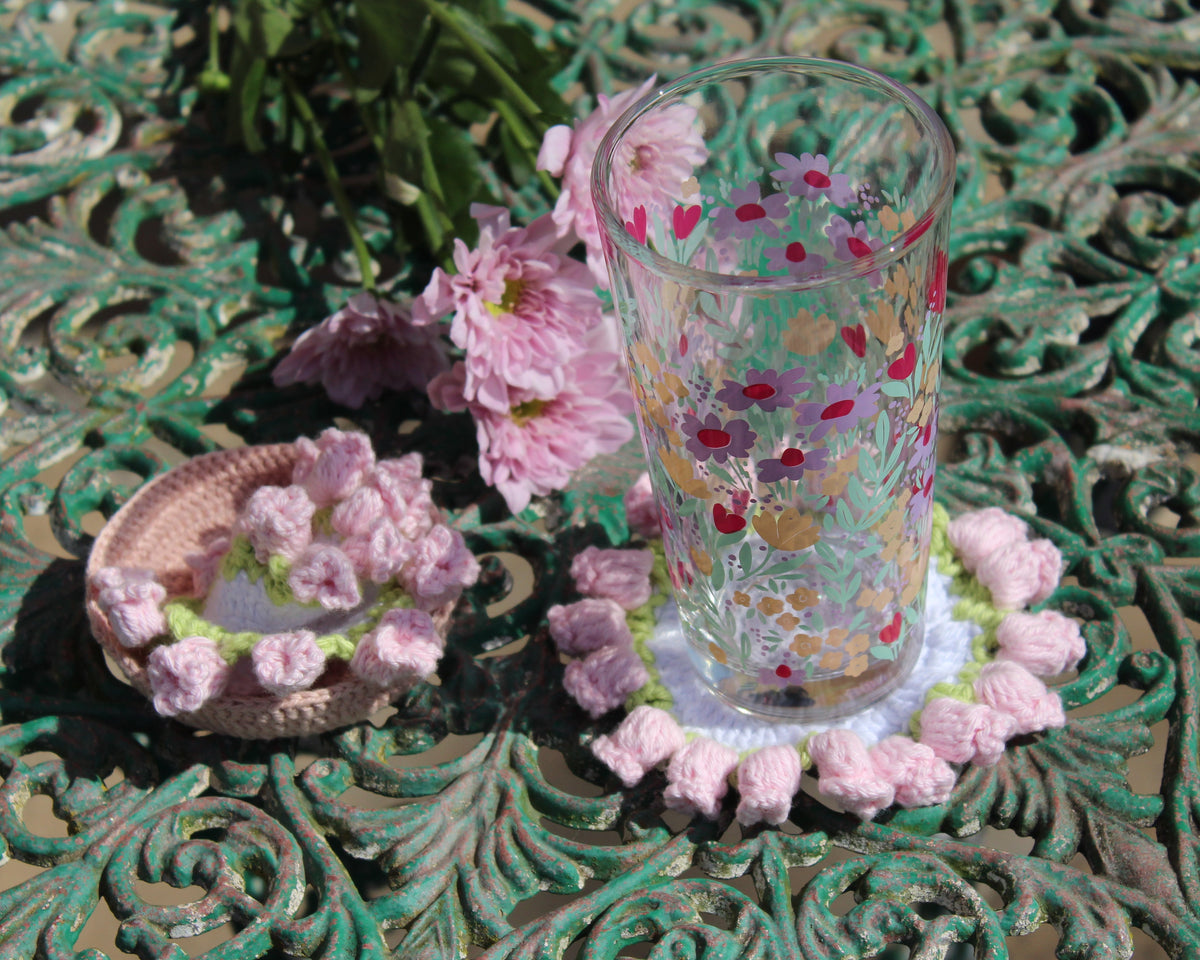 A clear glass tumbler decorated with pink and red flowers, resting on a pink crocheted coaster alongside a matching floral coaster lid, set on an ornate green metal surface with pink flowers in the background.