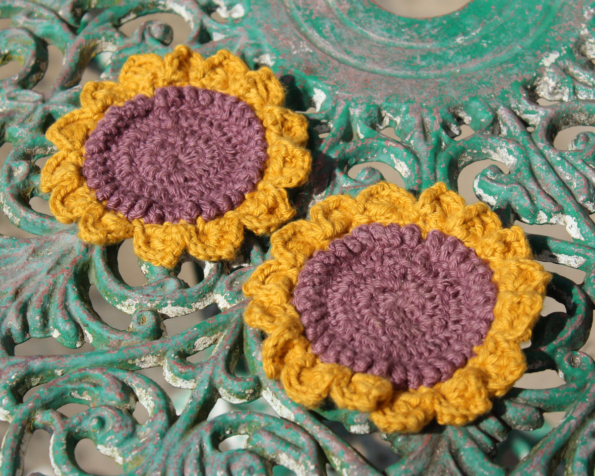 Two crocheted sunflower-shaped items with purple centers and mustard yellow petals, displayed on a distressed teal metal surface.